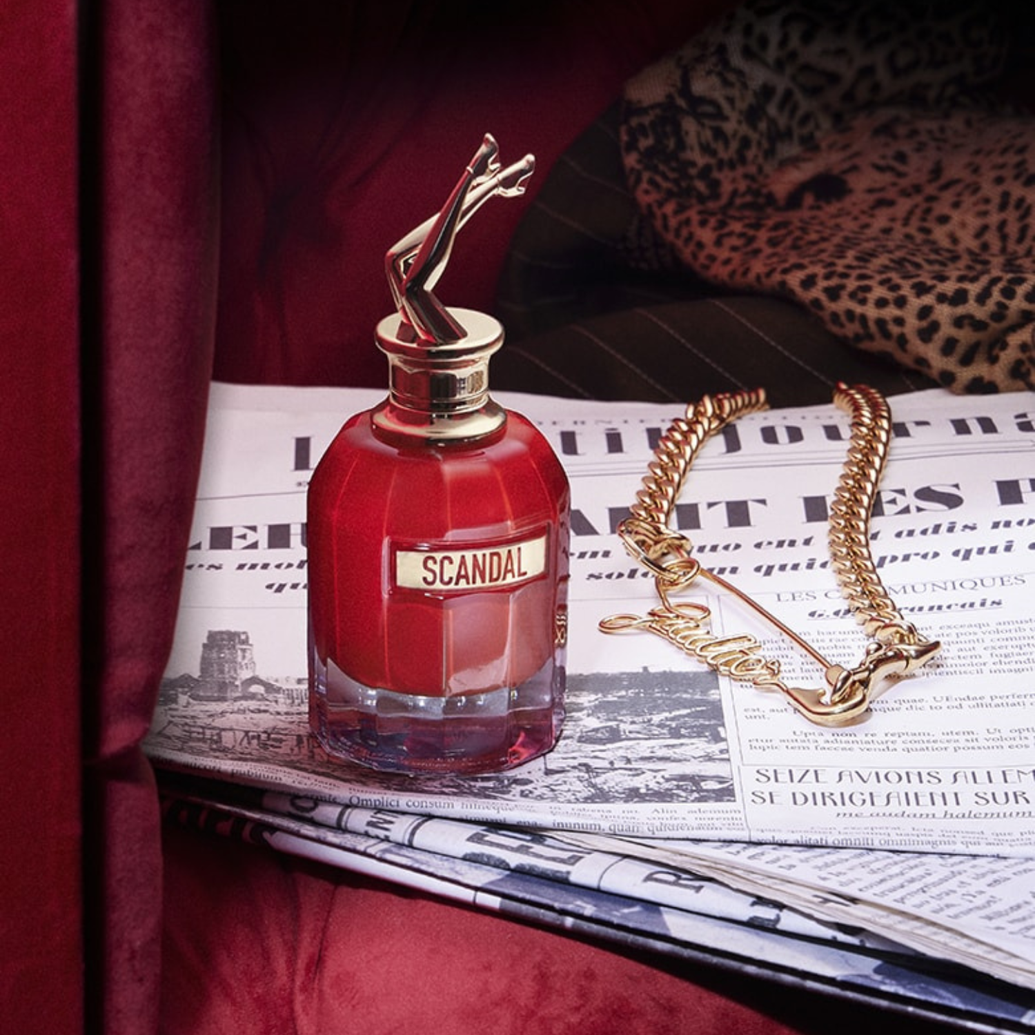 Perfume Scandal Le Parfum sobre jornal francês dobrado, ao lado de colar dourado com o nome "Gaultier", em cenário com tecido vermelho e estampa de oncinha ao fundo.