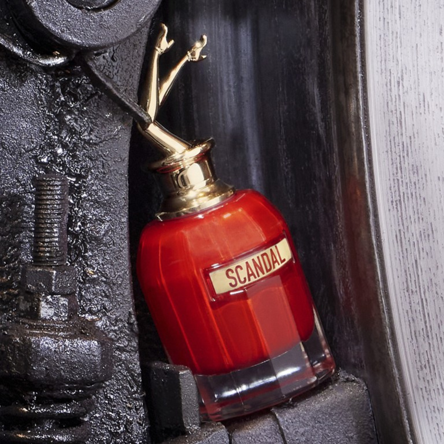 Frasco do Scandal Le Parfum encostado em uma estrutura metálica industrial, com contraste entre o brilho do vidro e da tampa dourada e a textura rústica do fundo.