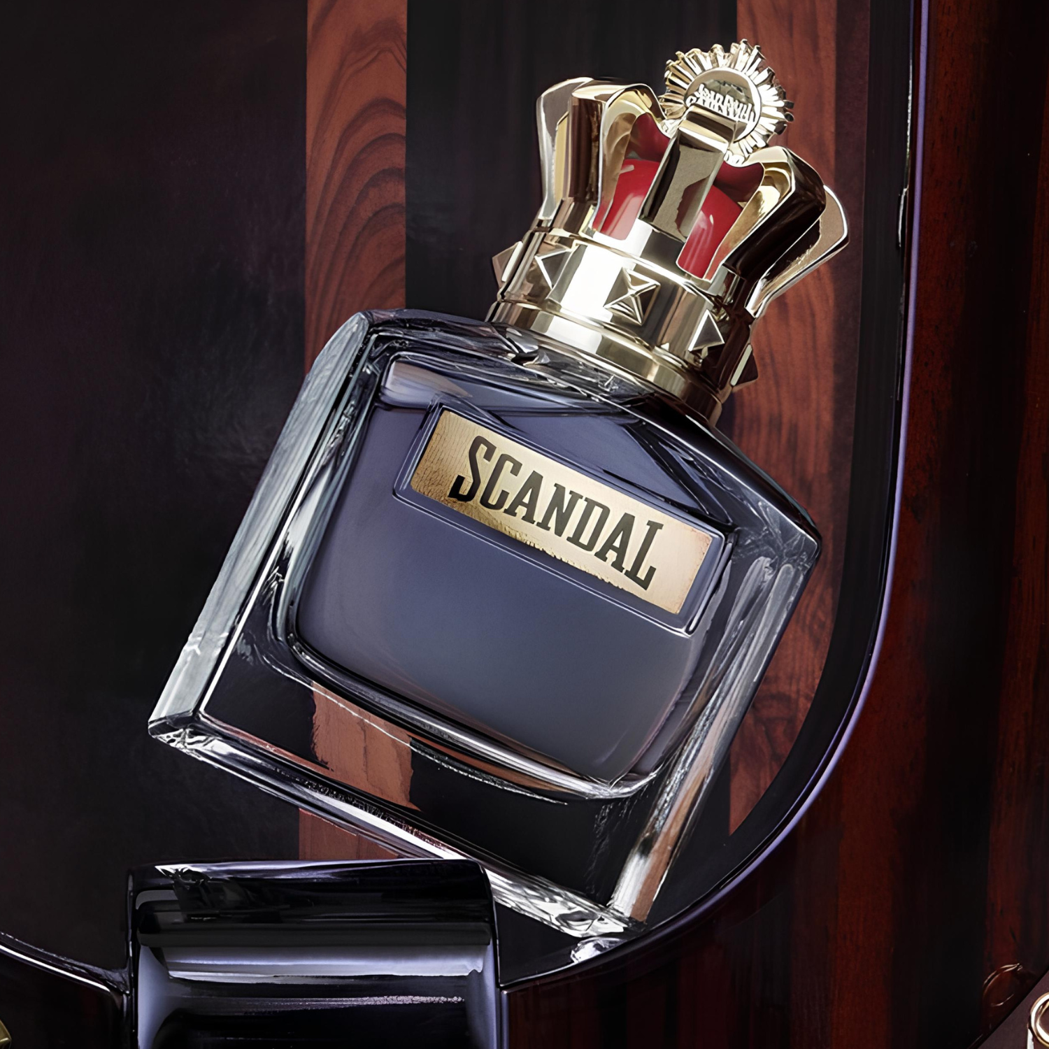 Perfume masculino Scandal Pour Homme Jean Paul Gaultier inclinado em uma base de madeira escura e brilhante, realçando o design da coroa dourada e o contraste com o líquido azul-cinzento.