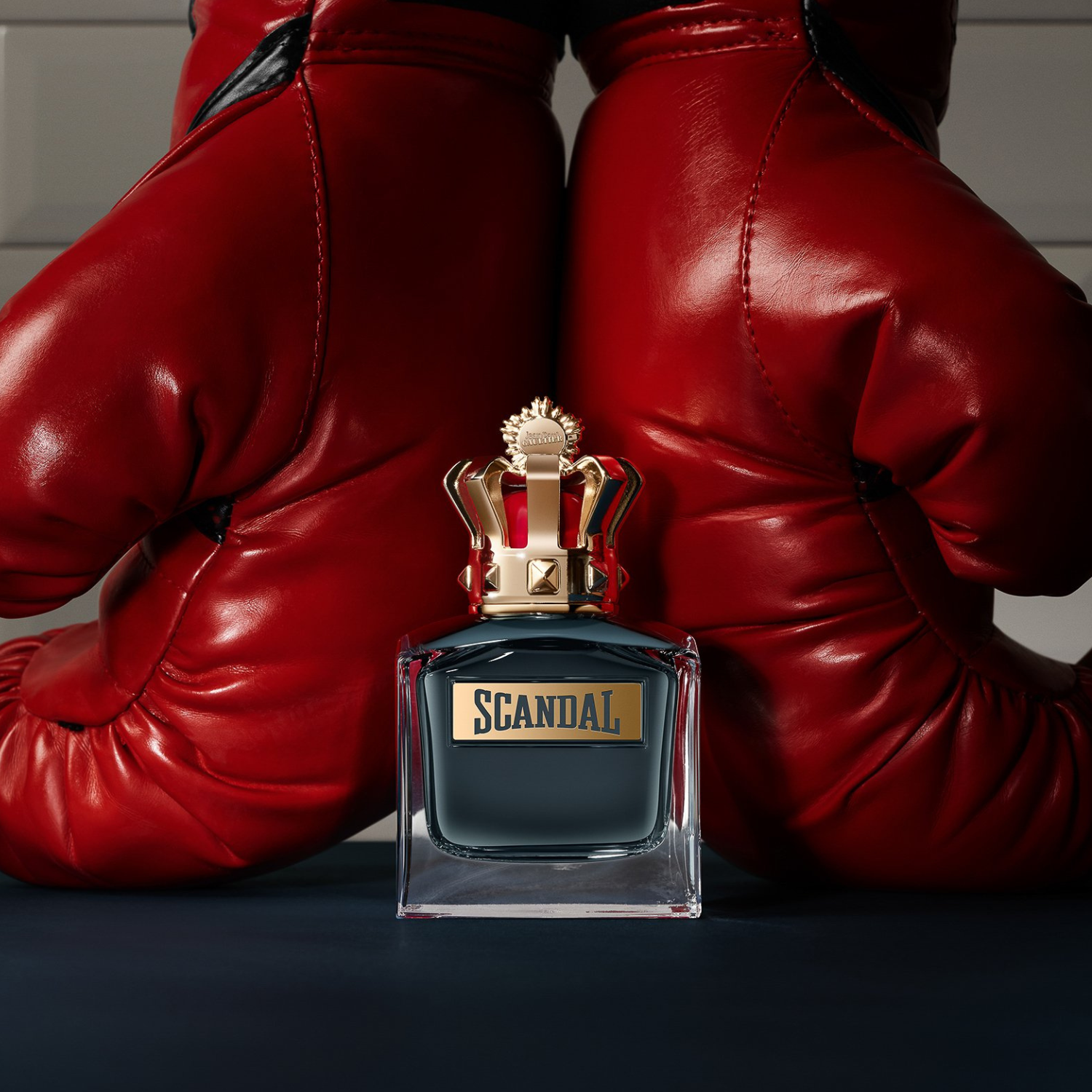 Perfume Scandal Pour Homme Jean Paul Gaultier em destaque em um ambiente com um par de luvas de boxe vermelhas ao fundo, transmitindo uma sensação de poder, luta e vitória.