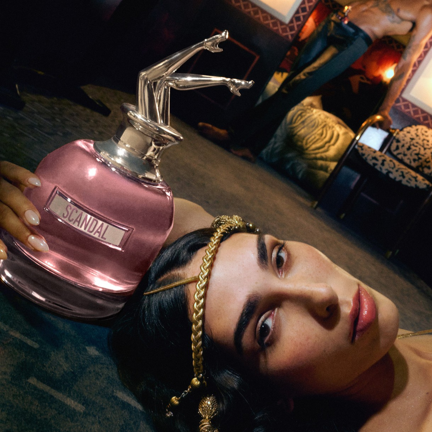 Mulher deitada segurando o perfume Scandal em destaque. Ela usa acessórios dourados, e o ambiente possui decoração luxuosa, ressaltando a atmosfera sedutora e audaciosa da fragrância.
