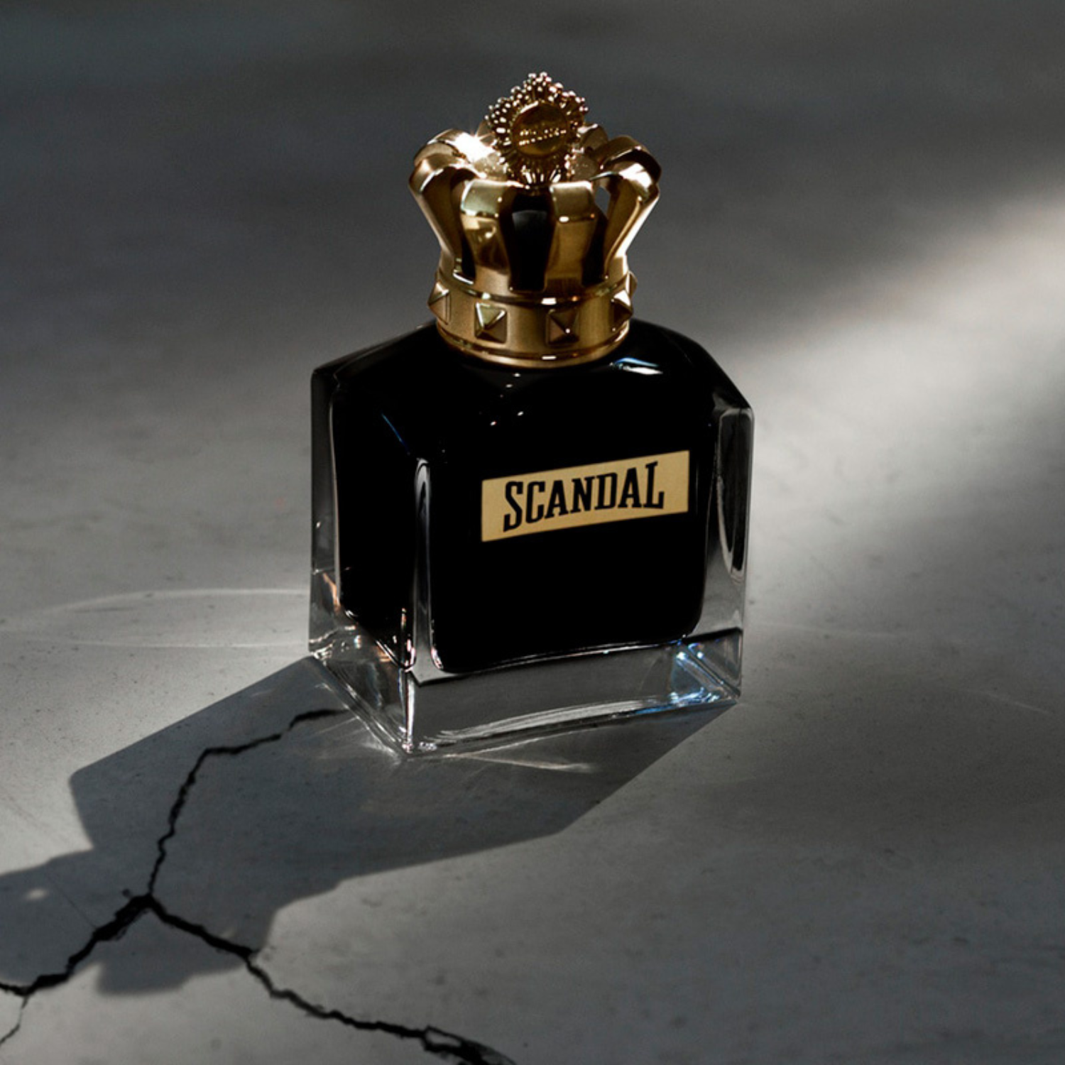 Frasco do perfume Scandal Pour Homme Parfum sob um feixe de luz em ambiente escuro, com a sombra do frasco projetada no chão rachado, transmitindo força e elegância.