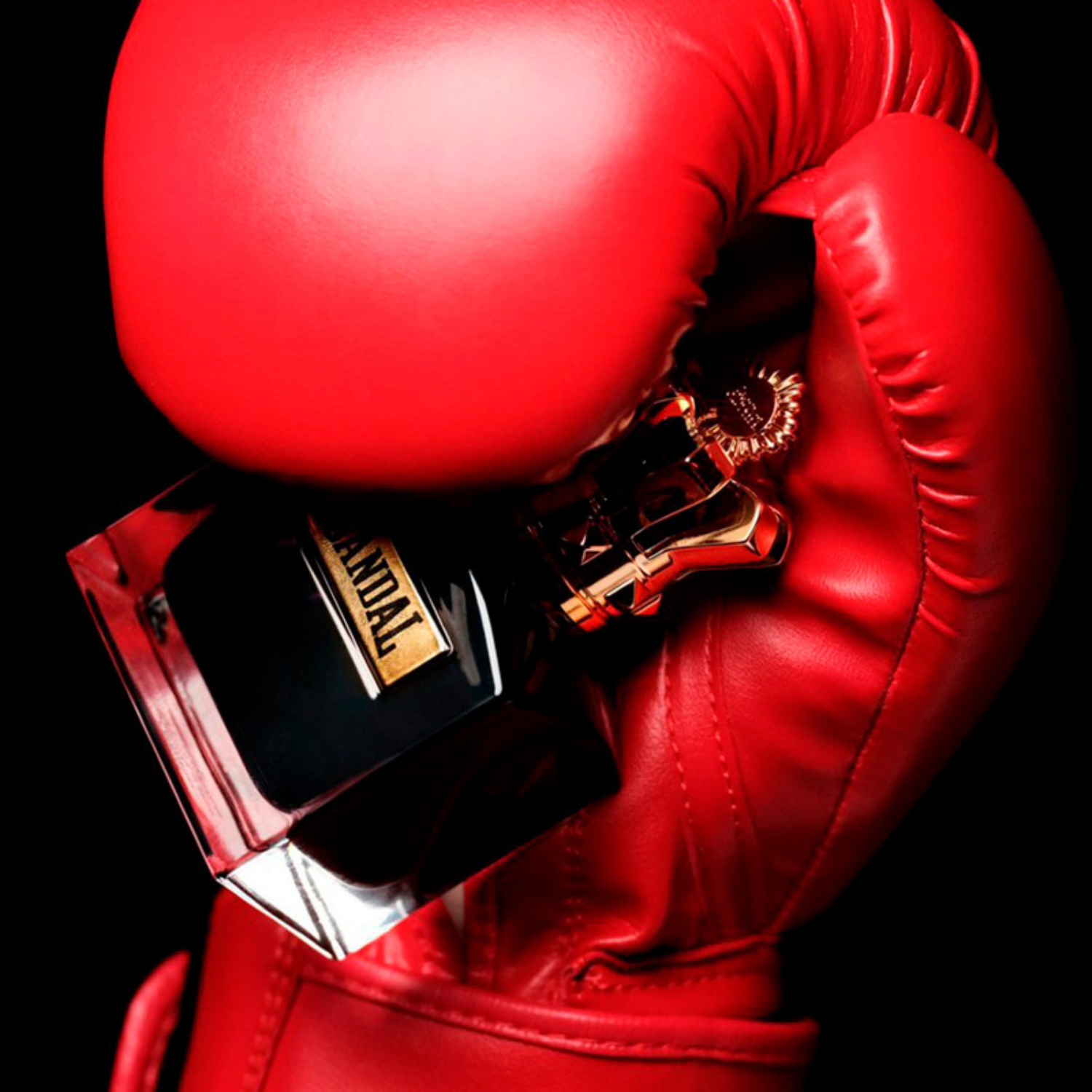 Frasco do Scandal Pour Homme Parfum segurado por uma luva de boxe vermelha, em contraste com o fundo preto, transmitindo intensidade, impacto e masculinidade poderosa.