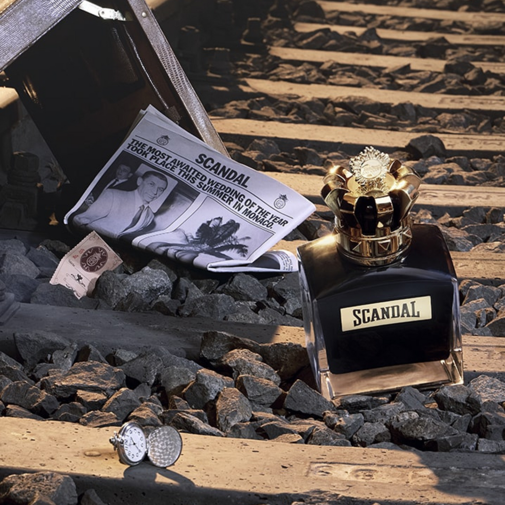 Frasco do Scandal Pour Homme Parfum posicionado sobre trilhos de trem, ao lado de um jornal fictício e acessórios masculinos, compondo uma cena dramática e cinematográfica que reforça o conceito ousado da fragrância.