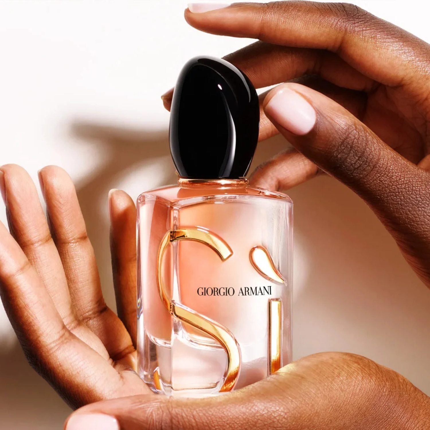 Mãos femininas de tons de pele diferentes segurando o frasco do perfume Sì Giorgio Armani, com unhas delicadas em tons claros, em um fundo suave, transmitindo uma sensação de cuidado e apreço pela fragrância.