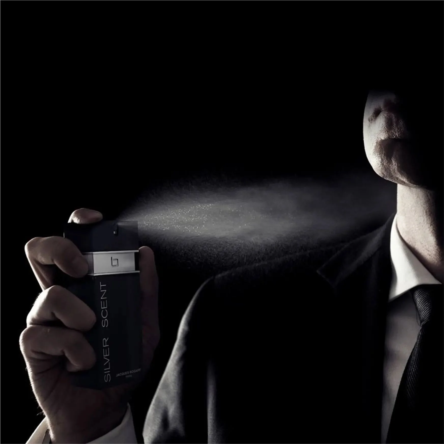 Homem de terno borrifando perfume Silver Scent Intense Jacques Bogart, com spray visível, sugerindo uso noturno e elegante da fragrância masculina.