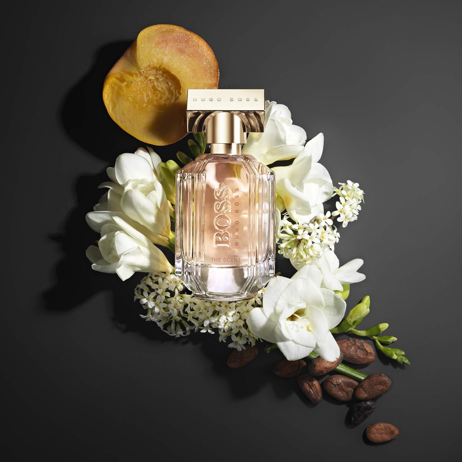 Perfume feminino The Scent for Her Boss em destaque em uma composição com uma metade de pêssego, flores brancas e grãos de cacau, sugerindo notas frutadas, florais e gourmand, com uma atmosfera sofisticada.
