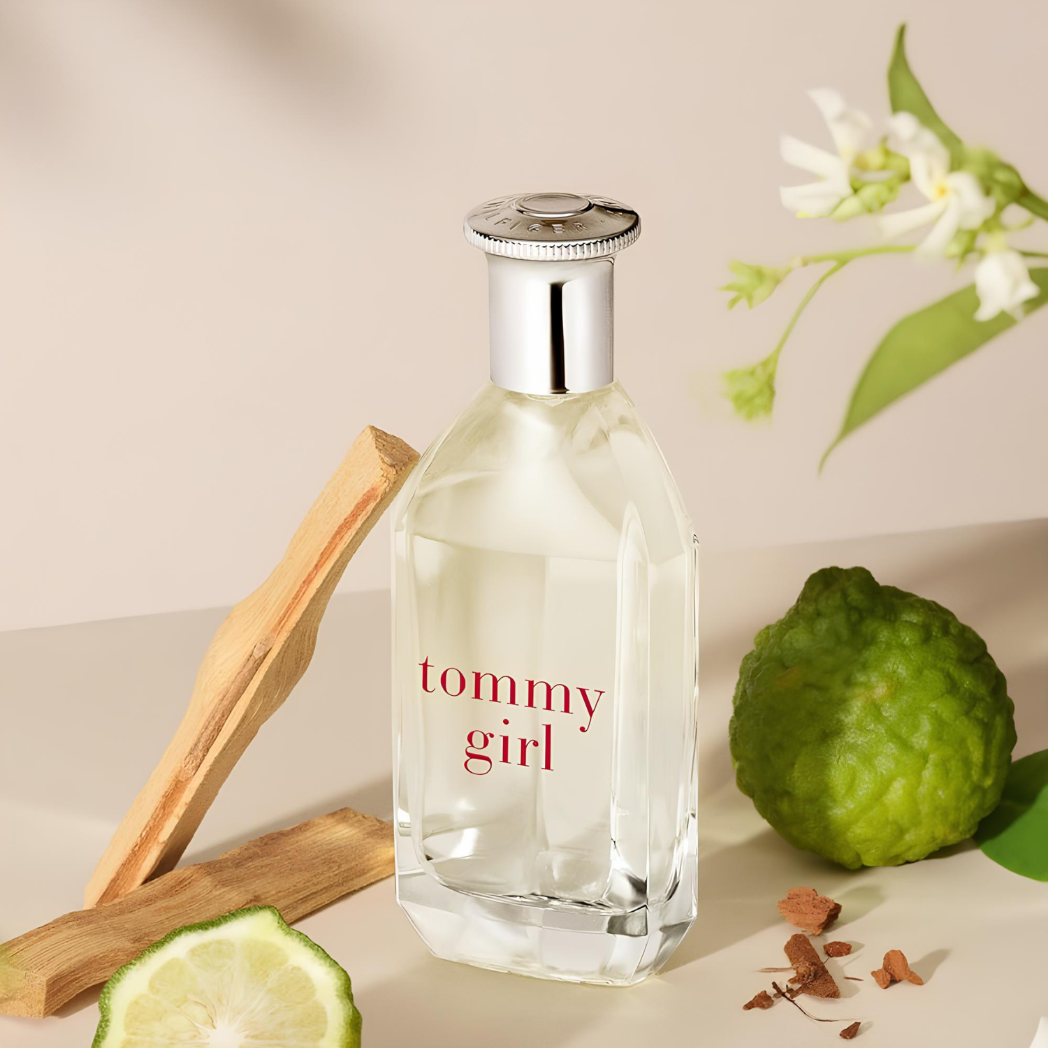 Perfume feminino Tommy Girl Tommy Hilfiger em destaque em uma composição com pedaços de madeira, um limão verde e flores brancas, sugerindo notas frescas, cítricas e naturais da fragrância.