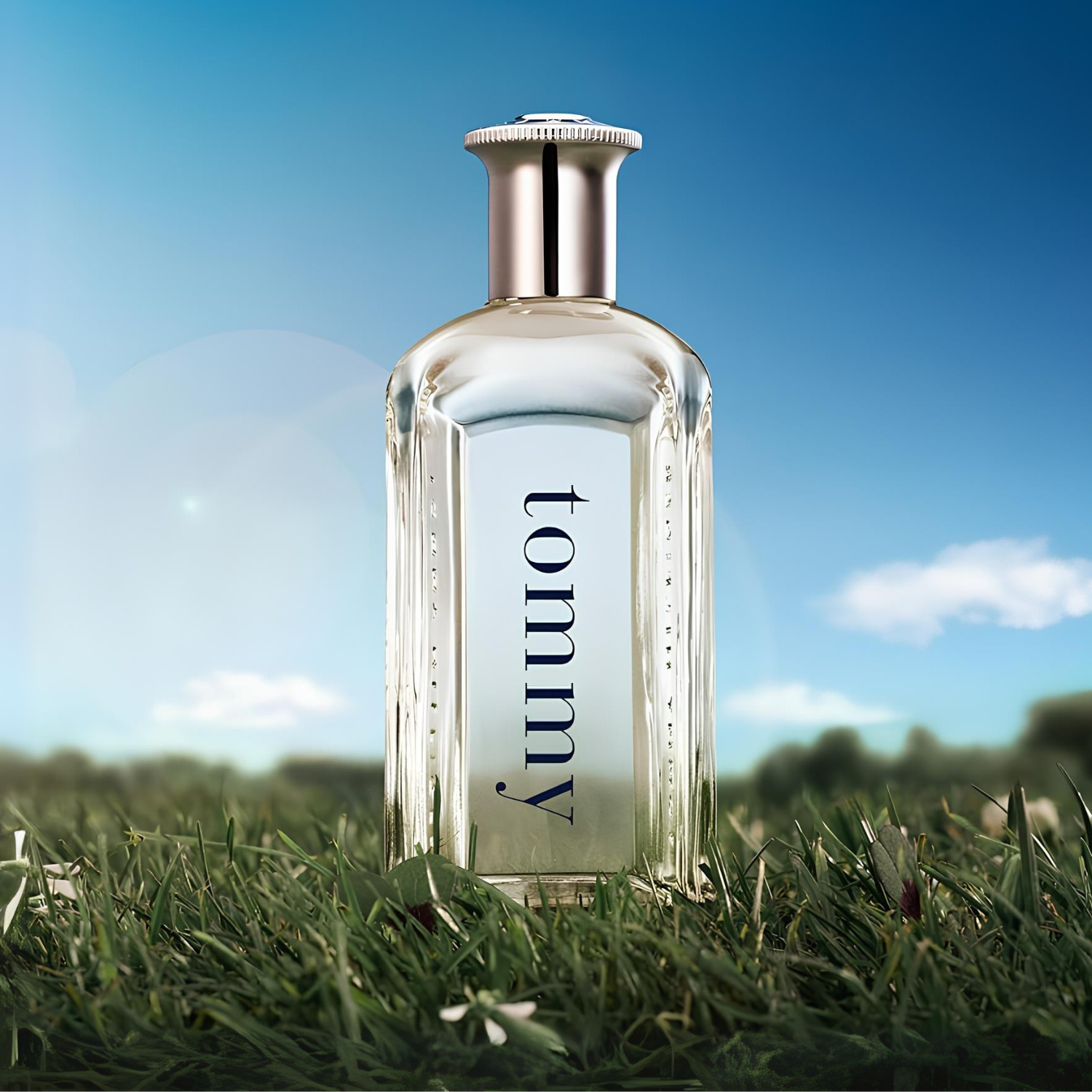 Perfume Tommy Men Tommy Hilfiger em destaque em um campo de grama verde sob um céu azul com nuvens, com uma luz suave e um fundo desfocado, transmitindo uma sensação de liberdade e frescor ao ar livre.