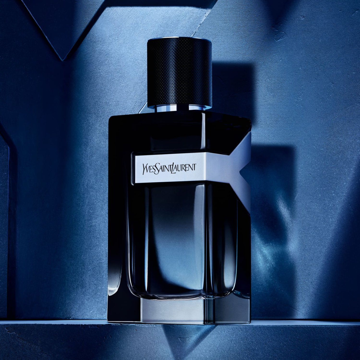 Perfume masculino Y Yves Saint Laurent em destaque em um ambiente com iluminação azul escura e sombras geométricas, realçando o design elegante e moderno do frasco.