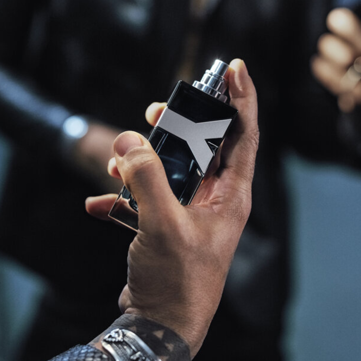 Close-up de uma mão masculina segurando o frasco do perfume Y Yves Saint Laurent, com foco no logo prateado "Y" e na embalagem sofisticada.